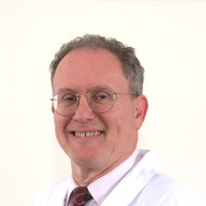 The image features a man wearing glasses, smiling at the camera, with gray hair, dressed in a white shirt and a tie, standing against a plain background.